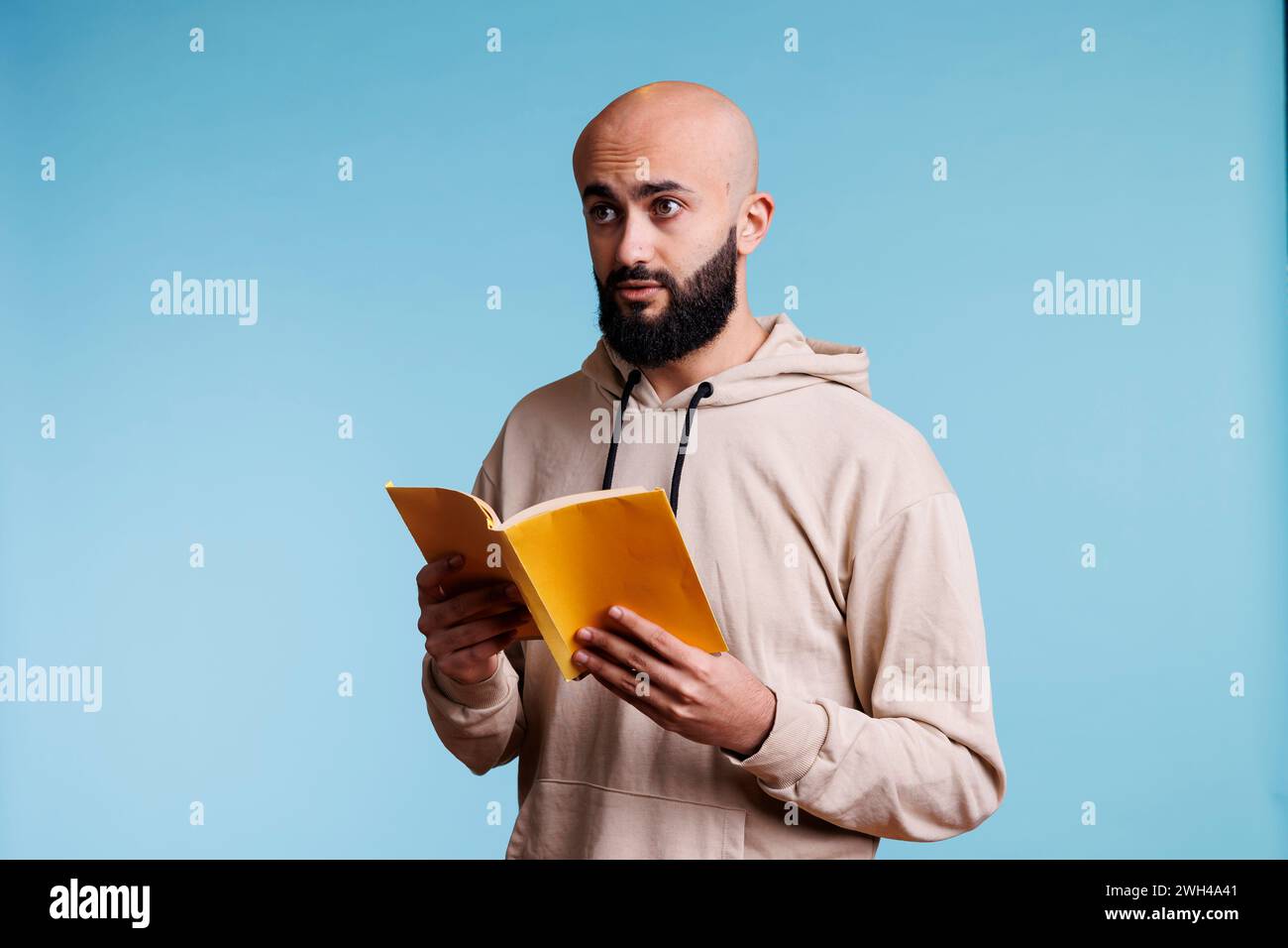Junger arabischer Mann, der ein Softcover-Buch hält und über einen Plan nachdenkt, während er wegblickt. Arabische Person liest Roman Taschenbuch mit gelbem Cover, während sie mit nachdenklichem Ausdruck steht Stockfoto