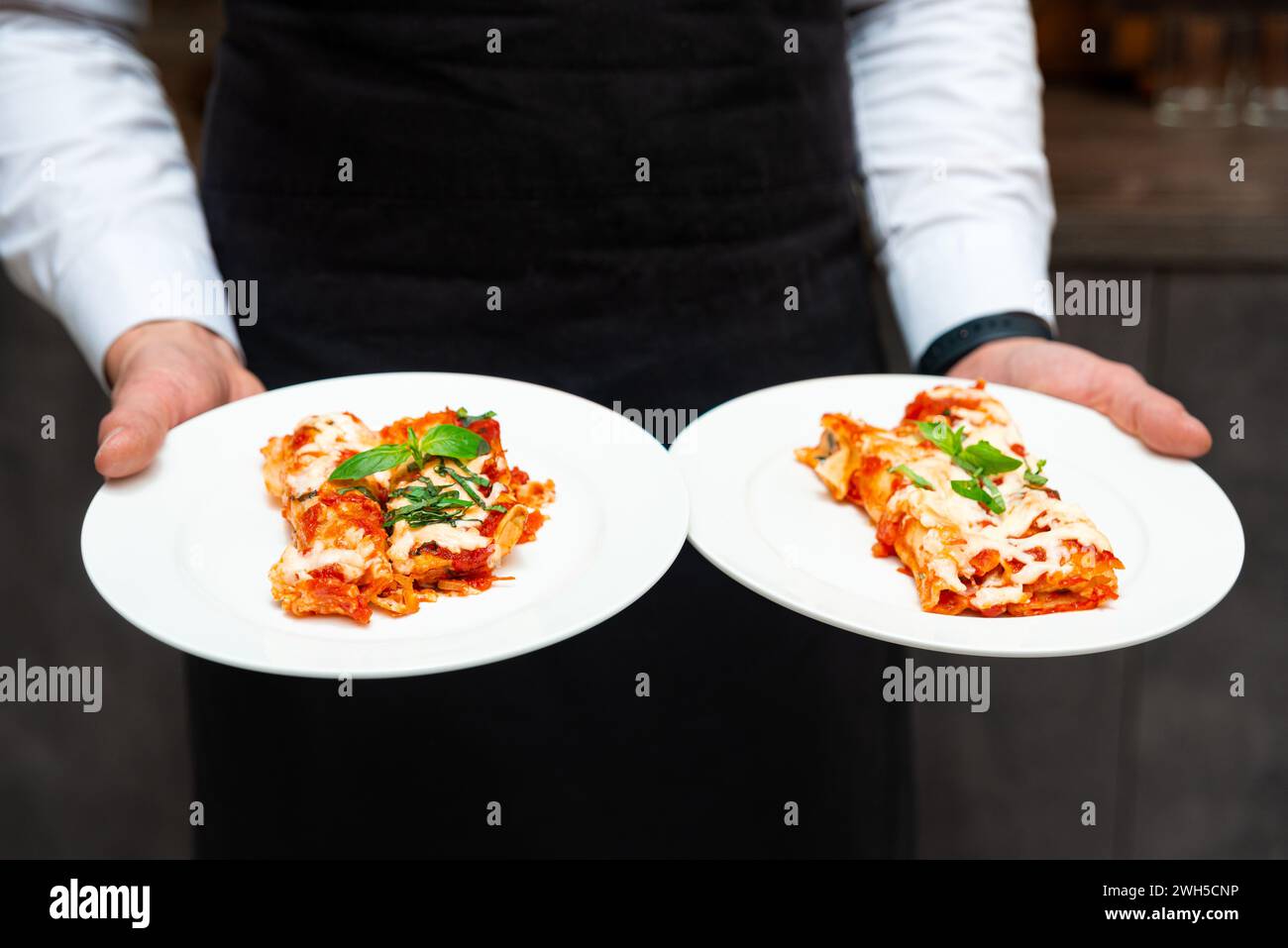 Ein professioneller Kellner präsentiert zwei Teller mit köstlicher Lasagne, garniert mit Basilikum, und präsentiert hochwertige italienische Küche im Restaurant. Stockfoto