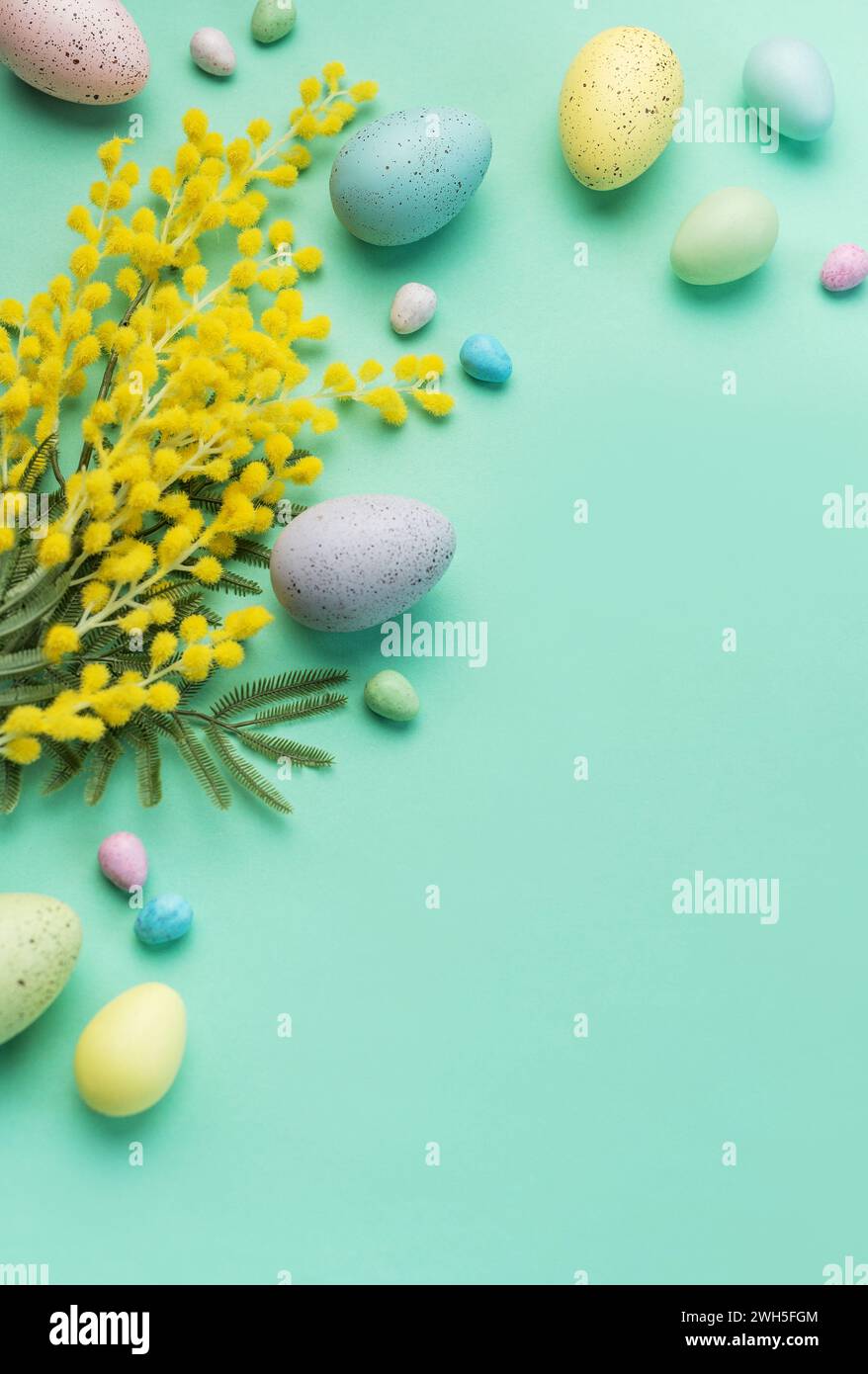 Ein kreatives Arrangement aus pastellfarbenen Ostereiern, Mimosa-Zweigen und Bonbons auf einer weichen grünen Oberfläche, das die Frühlingsfeier symbolisiert Stockfoto