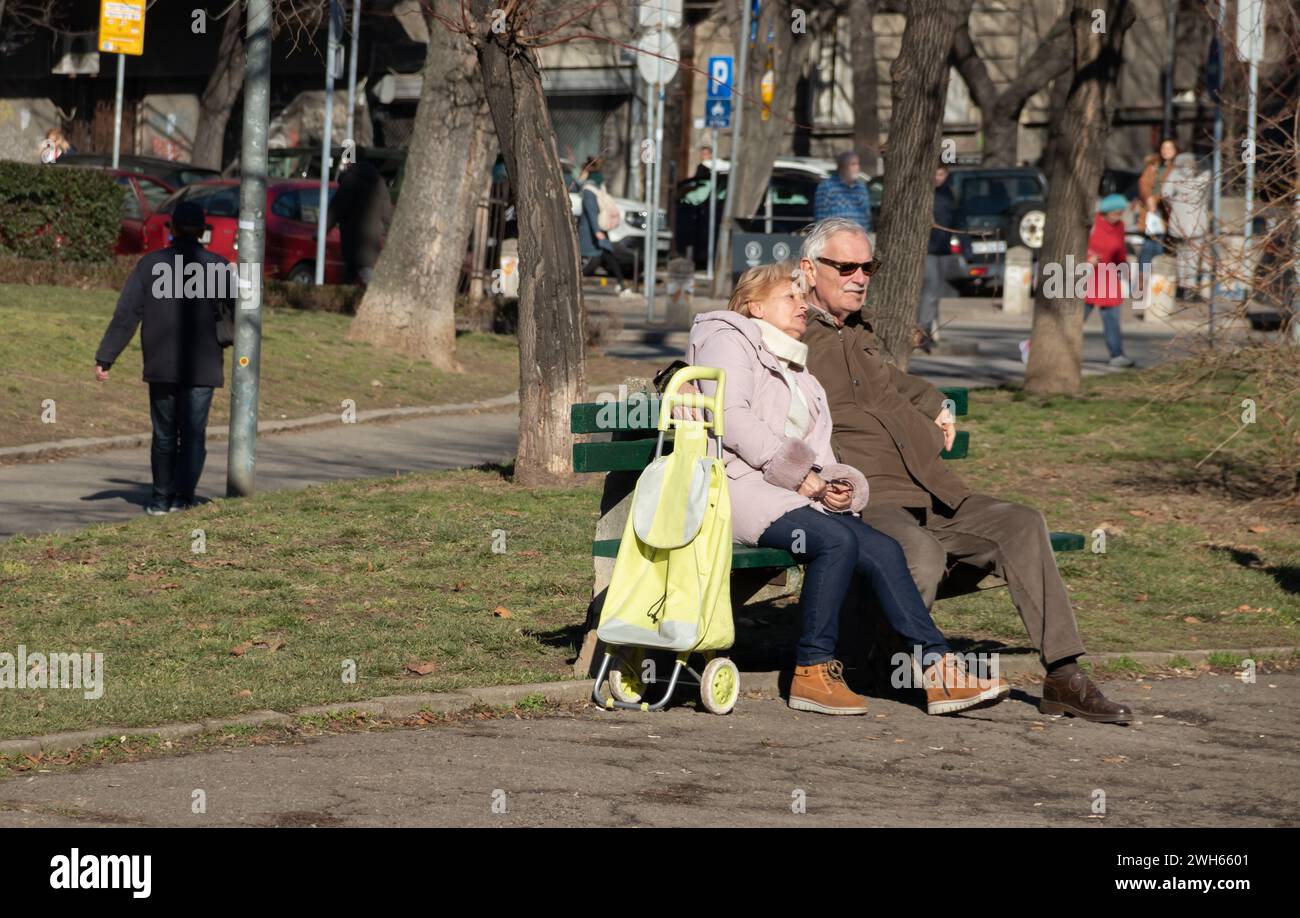 Ein altes Paar sitzt auf einer Bank in einem öffentlichen Park, mit ihr, die sich auf ihn stützt, und spiegelt ihre Liebe, Gesellschaft und ihren zärtlichen Moment wider Stockfoto