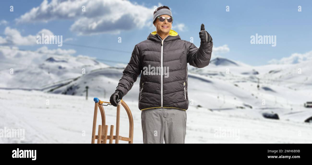 Mann auf einem schneebedeckten Berg mit Holzschlitten und Daumen hoch Stockfoto