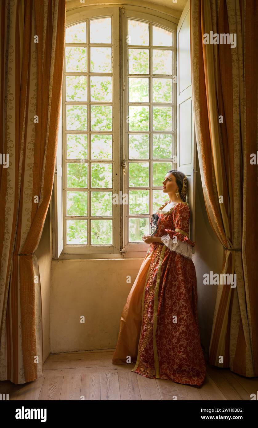 Frau mit authentischem Renaissance-Kostüm und Kopfschmuck, die in einem mittelalterlichen Schloss an ihrem Fenster steht Stockfoto