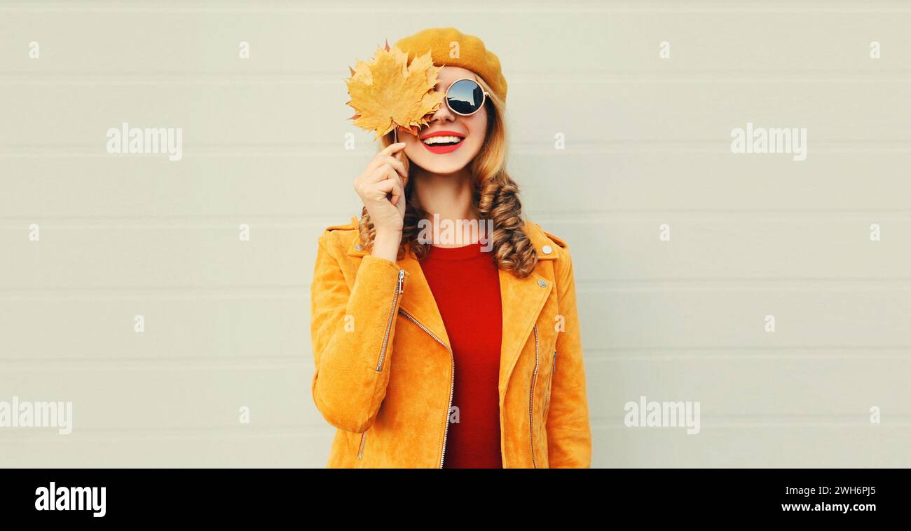 Herbstporträt der schönen glücklichen lächelnden jungen Frau mit gelben Ahornblättern, die einen französischen Baskenmütze, Sonnenbrille und Jacke auf grauem Hintergrund trägt Stockfoto