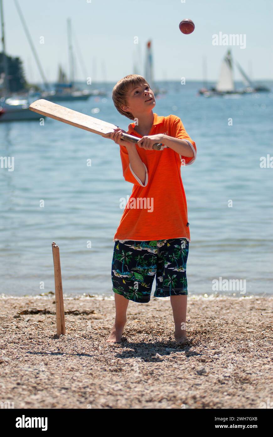 Ein kleiner Junge spielt während der Sommerferien Beach Cricket. Er reiht den Ball auf, um ihn für einen Lauf zu treffen. Stockfoto