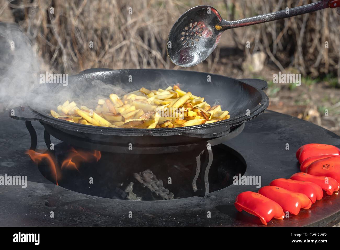 Gemüse, Paprika und Kartoffeln werden an einem runden Kamin im Freien gebraten. Runder Grill in Form einer Schüssel mit Feuer im Inneren. Rundes Metallholz Stockfoto