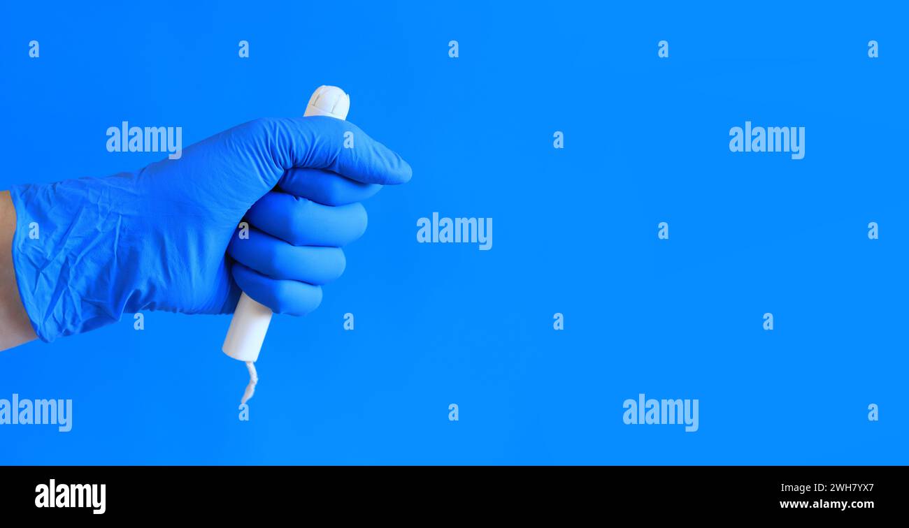 Tampon in einem Pappappappapplikator in der Hand einer Frau, blauer Hintergrund. Eine Hand in einem blauen medizinischen Latexhandschuh hält einen Tampon. Weibliche Körperhygiene PR Stockfoto