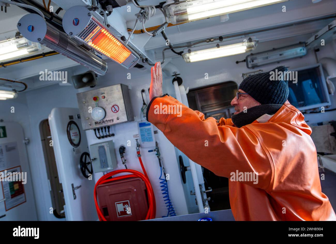 Sassnitz, Deutschland. Februar 2024. Marko Weißköppel wärmt sich zwischen dem Einsatz einer Hydrografiesonde an Bord des Fischereiforschungsschiffs Clupea in der Ostsee vor Rügen die Hände an einem Heizstrahler. Die Sonde dient zur Messung des Sauerstoffgehalts, der Wassertemperatur und des Salzgehalts in allen Wassertiefen, die später analysiert werden. Wissenschaftler des Thünen-Instituts untersuchen derzeit die Auswirkungen der Bauarbeiten für die Erdgaspipeline von Mukran nach Lubmin. Große Heringsschwärme versammeln sich im Winter in der Sassnitzer Gully und wandern ab Credit: dpa/Alamy Live News Stockfoto