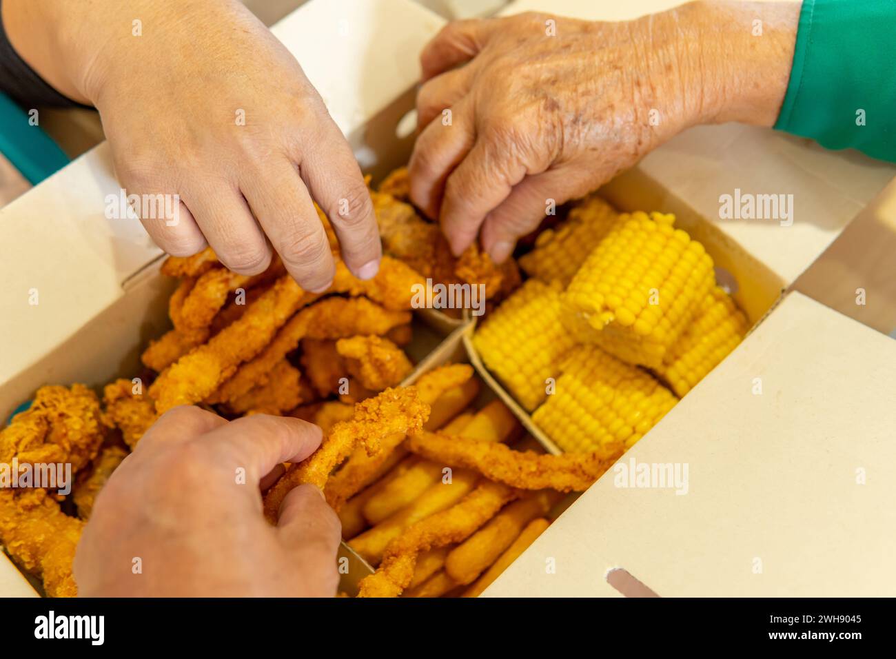 Nahaufnahme von drei Händen, die Essen aus einer Fast-Food-Box mit Huhn, Mais, Pommes frites und Arepas nehmen Stockfoto