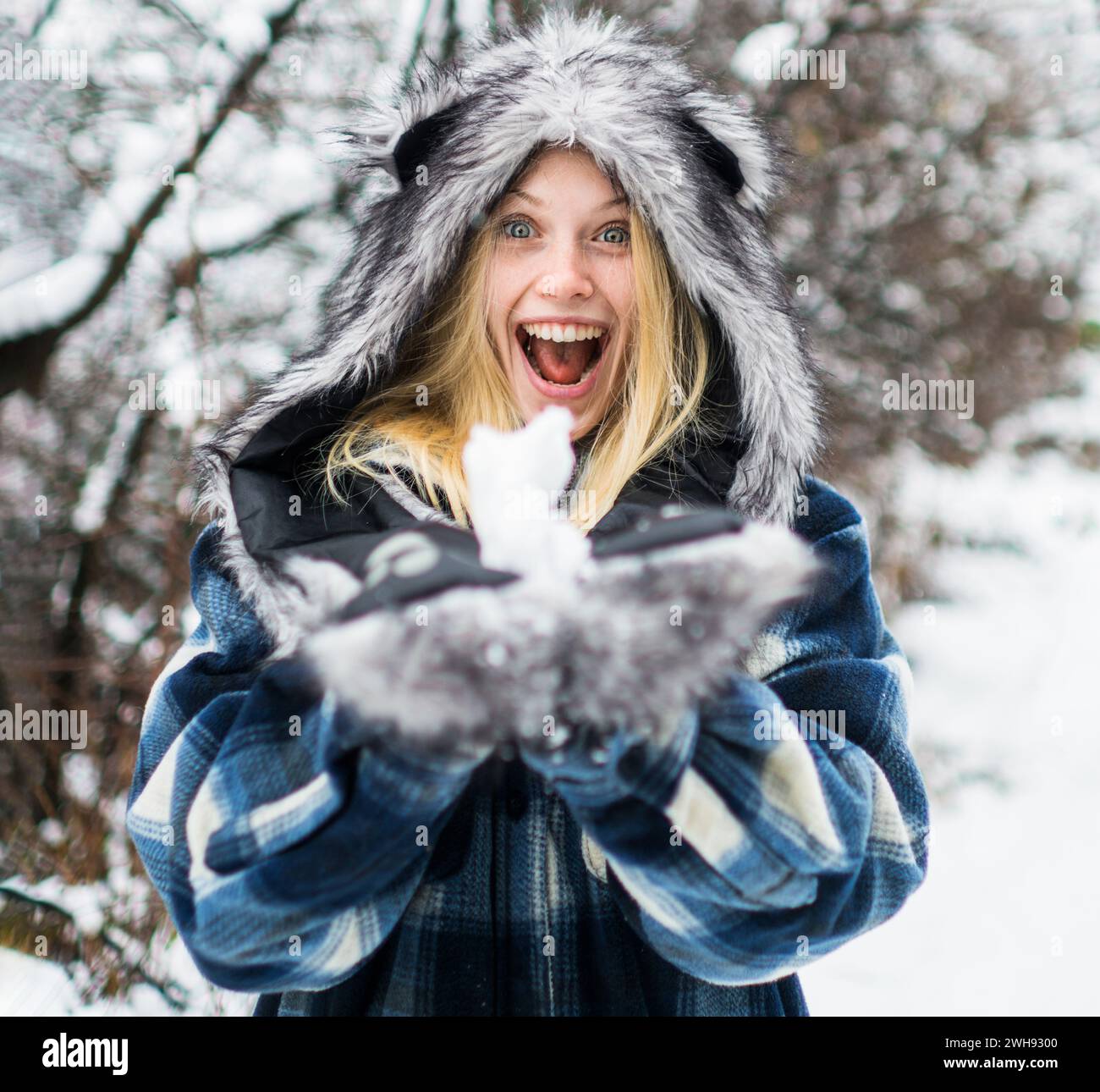 Winterzeit. Wintermädchen spielt mit Schnee im Park. Glückliche Frau in warmer Kleidung mit Schneebälle. Schneebedecktes Wetter. Blondes Mädchen in kariertem Mantel, Pelzmütze und Stockfoto