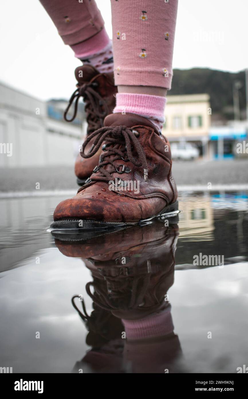 Nahaufnahme eines Mädchens in rosa Strumpfhosen mit nassen braunen Stiefeln, das sich in einer Regenpfütze auf der Straße spiegelt. Stockfoto
