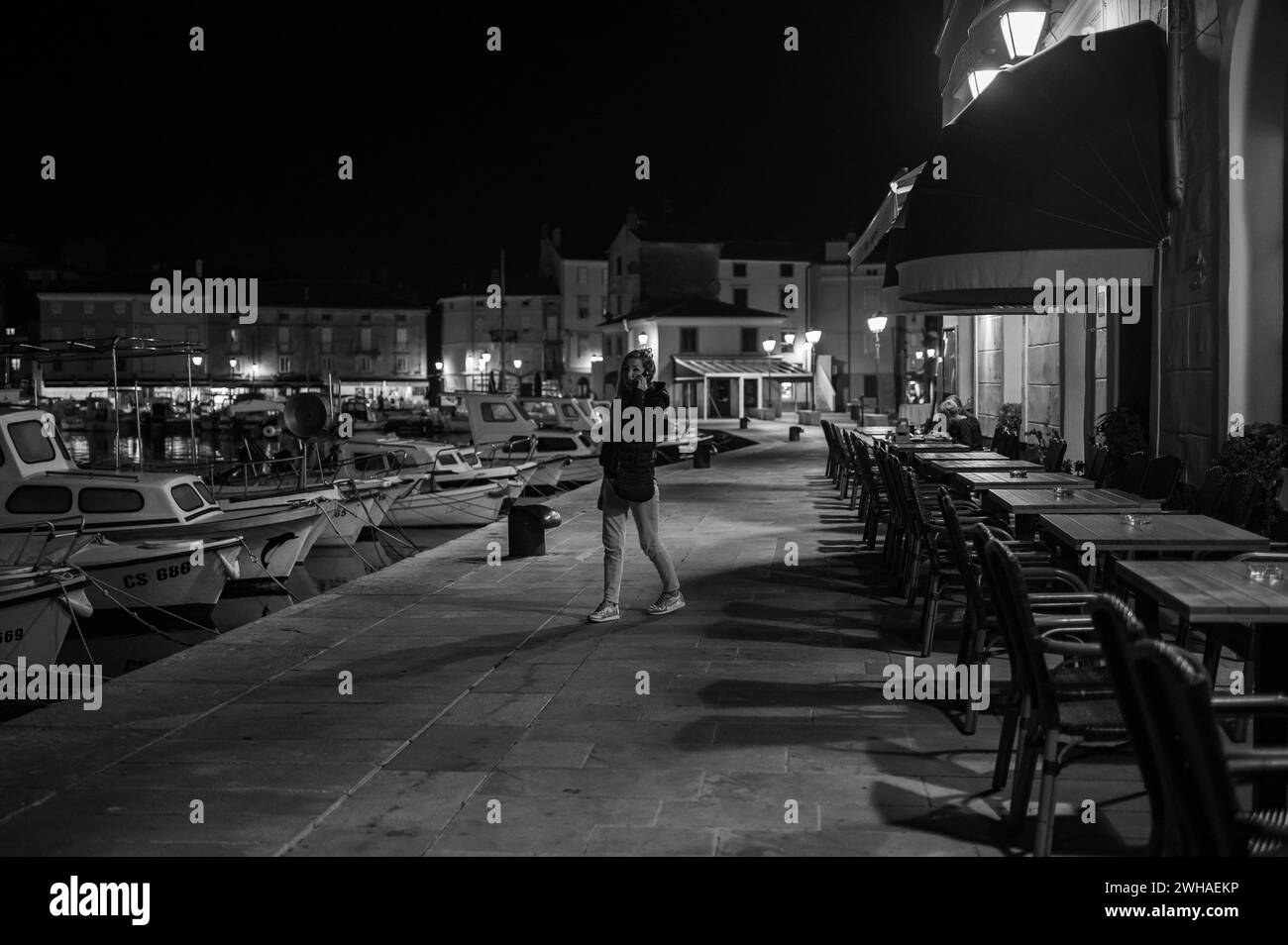 Cres, Kroatien - 24. Oktober 2022: Der Hafen von Cres in der Nacht, leere Stühle einer Bar, Frau am Telefon, schwarz-weiß Stockfoto