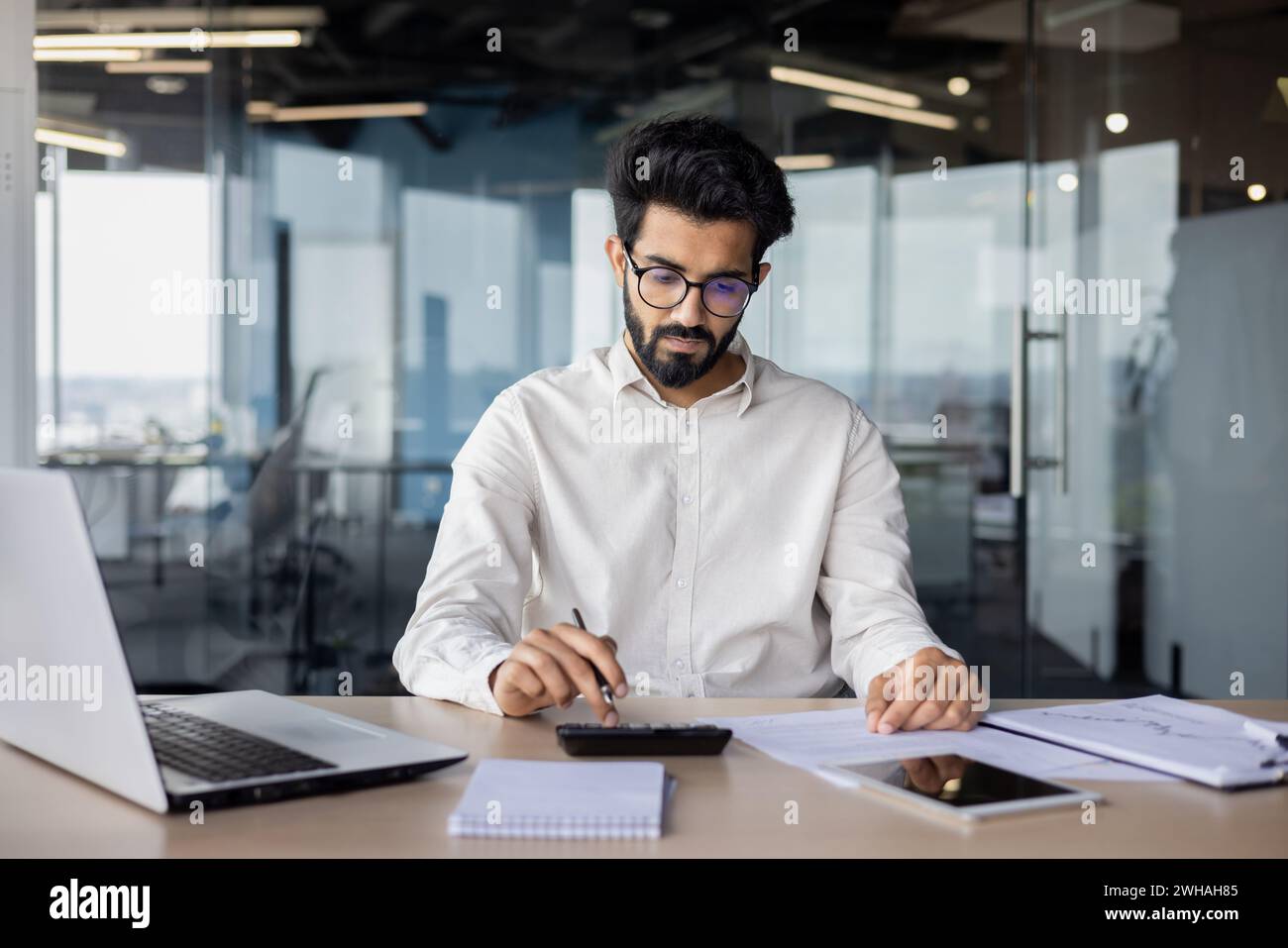 Ein indischer Geschäftsmann mit Bart arbeitet intensiv an seinem Laptop, umgeben von einem modernen Büroinnenraum, das für engagierte Professionalität steht. Stockfoto
