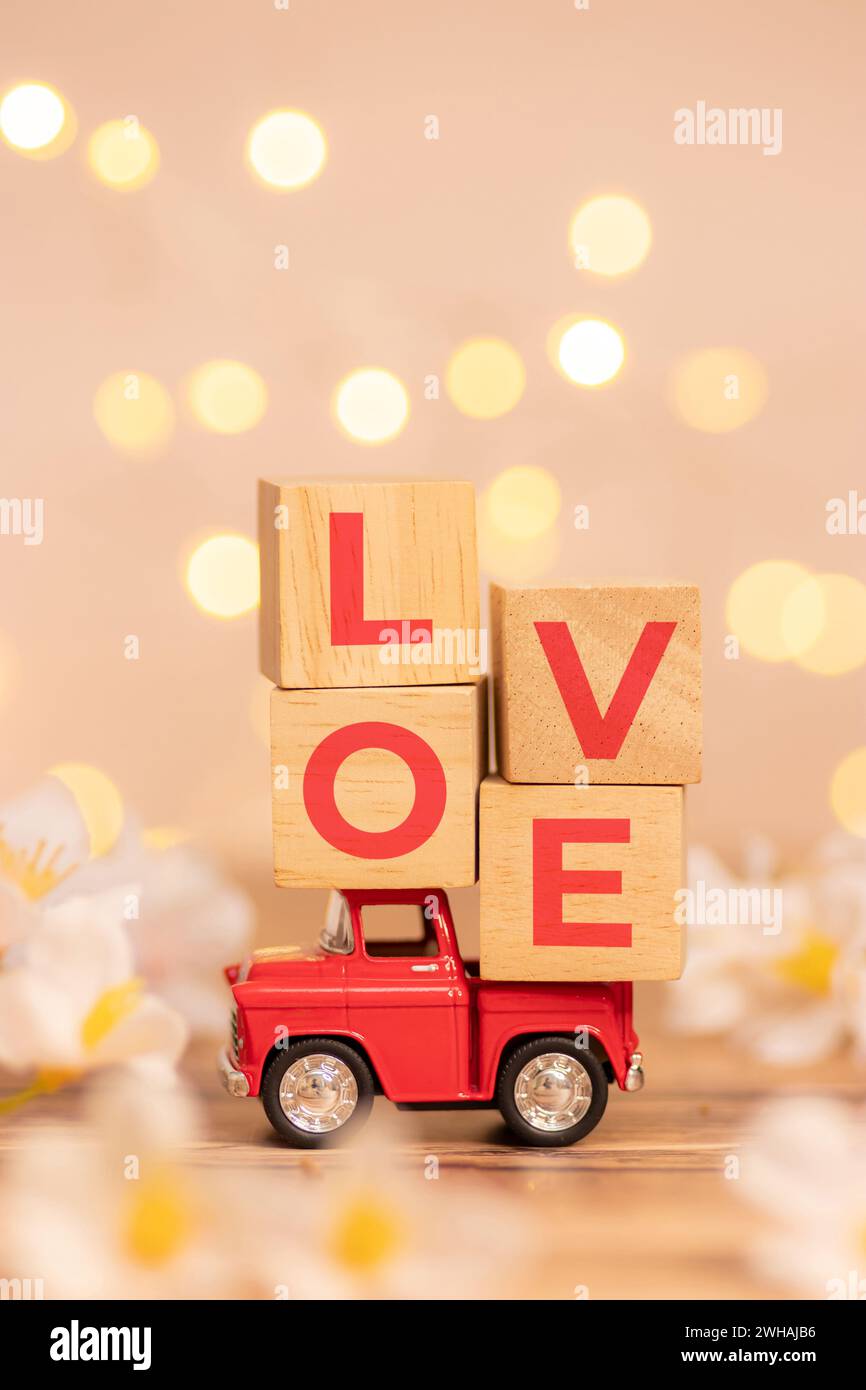 Rotes Truck-Spielzeug mit Holzwürfel und Text Love auf der Rückseite des Pickups, um Liebe zum Valentinstag mit unscharfem weißem Blumenfeld und d zu liefern Stockfoto
