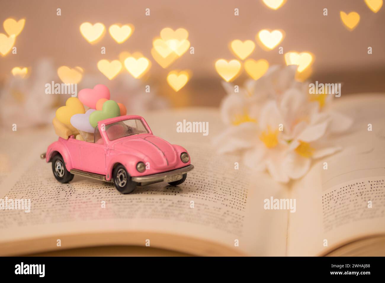 Ein rosafarbenes Oldtimer-Cabriolet trägt ein farbenfrohes Herz im Inneren, um Liebe zum Valentinstag zu liefern, auf einem unscharfen Roman mit ausgebreiteten Seiten, defokussiertem Herz Stockfoto