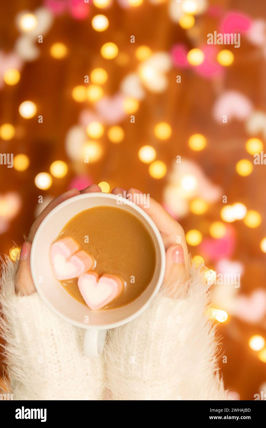 Frauenhand mit Pullover, der eine Kaffeetasse mit Marshmallow in Herzform hält, defokussiertes Bokeh-Licht für Valentinstag-Feier dekoriert Stockfoto