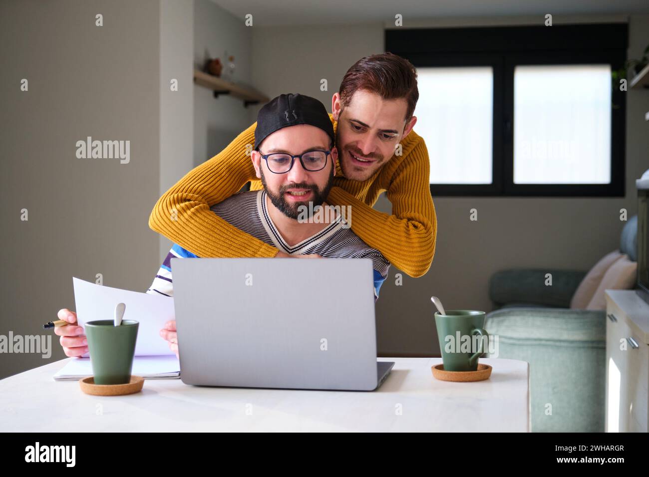 Homosexuelles Paar, das zu Hause an einem Laptop arbeitet und umarmt. Stockfoto