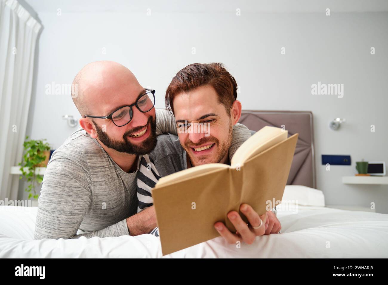 Homosexuelles Paar liest ein Buch, umarmt und lacht zusammen im Bett. Stockfoto