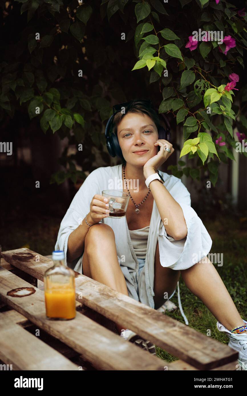 Frau in Kopfhörern trinkt Kaffee, Café, sitzt auf dem Rasen. Bali. Stockfoto