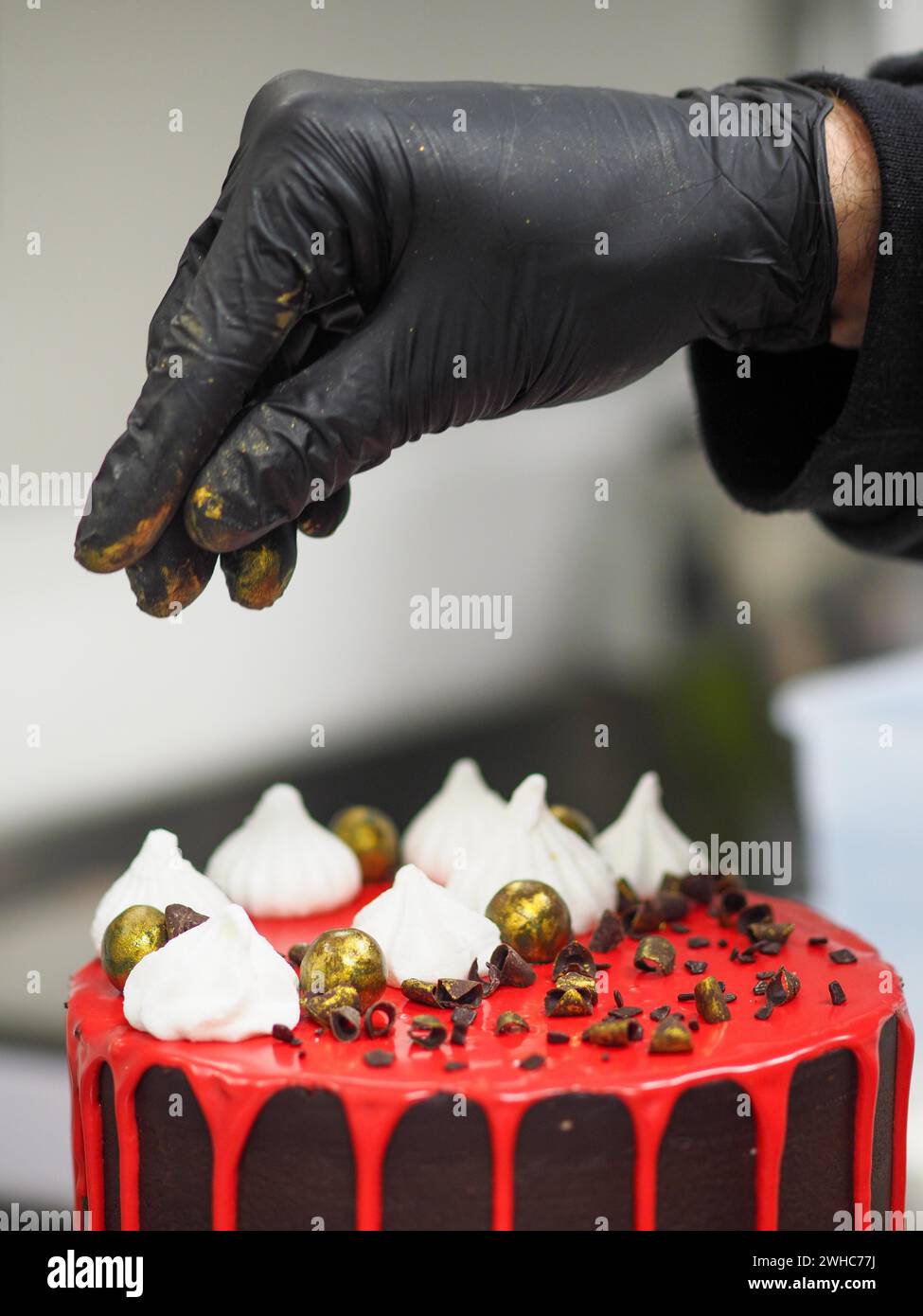 Ein professioneller Konditor trägt schwarze Handschuhe, die einen Schokoladenkuchen kunstvoll mit goldbemalten Schokoladenstreuseln dekorieren Stockfoto