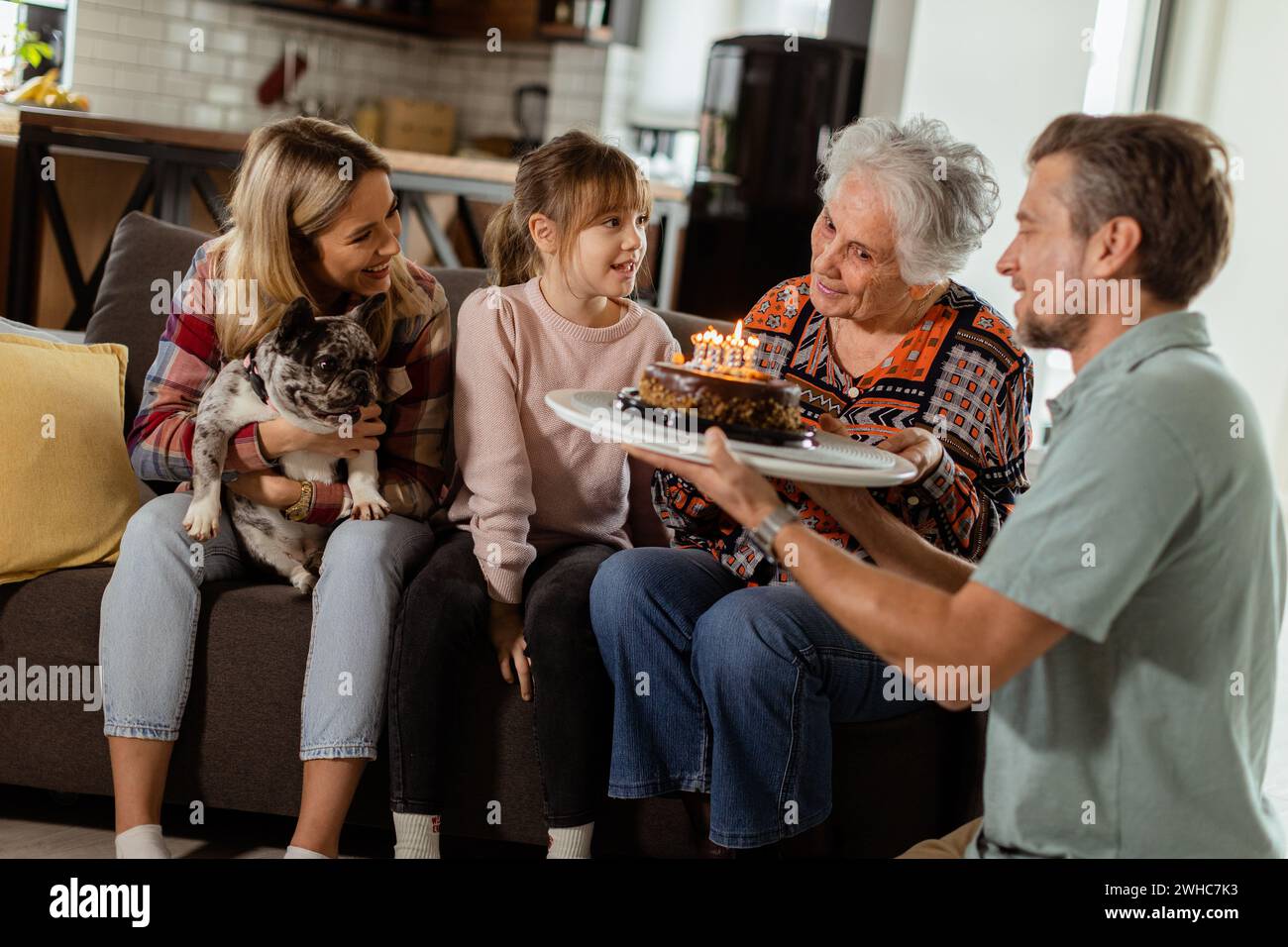 Eine herzerwärmende Szene entfaltet sich, während sich eine Familie mit mehreren Generationen auf einer Couch versammelt, um einer entzückten Großmutter einen Geburtstagskuchen zu präsentieren und Erinnerungen zu schaffen Stockfoto