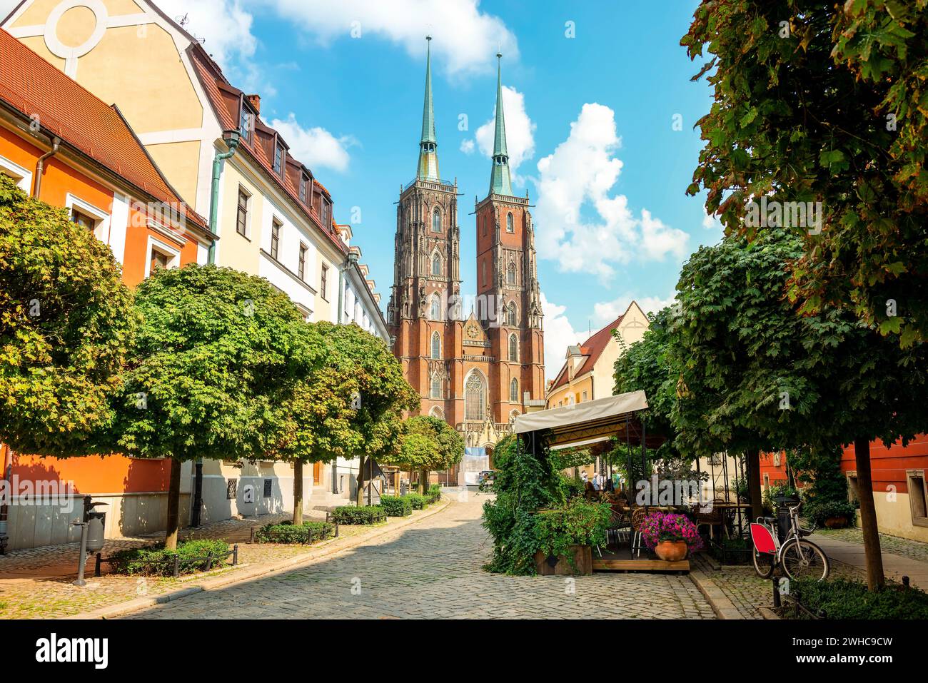 Straße mit Kopfsteinpflasterstraße mit grünen Bäumen, farbenfrohen Gebäuden, Sommercafe, Kathedrale von St. Johannes-der-Täufer-Kirche mit zwei Türmen im alten historischen Stadtzentrum, Ostrow Tumski, Breslau, Polen Stockfoto