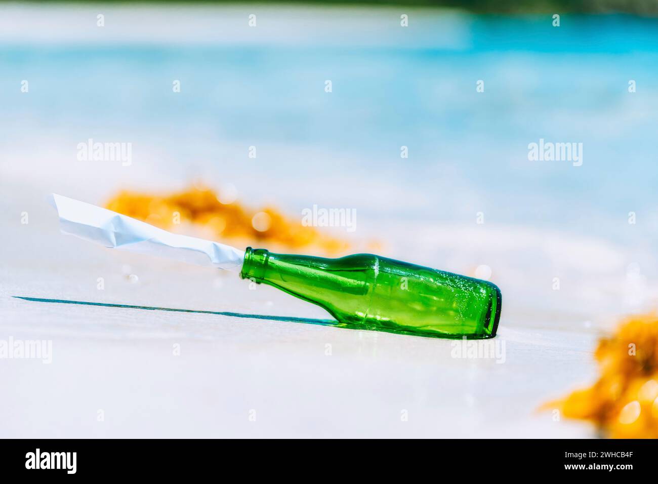 Grüne Flasche mit Papiernachricht auf weißem Sandstrand mit blauer Lagune im Hintergrund. Stockfoto