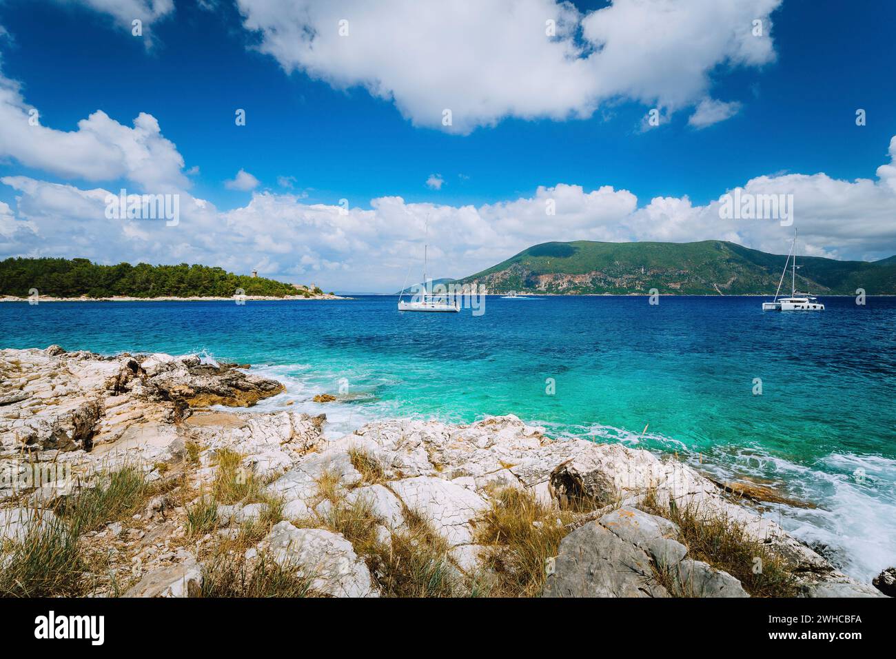 Yachten treiben mitten im offenen kristallklaren, smaragdgrünen Meerwasser in der Nähe von Fiskardo. Ithaki Island im Hintergrund. Erstaunliche weiße Wolkenlandschaft, die sich im blauen Himmel bewegt, Kefalonia, Ionische Inseln, Griechenland. Stockfoto