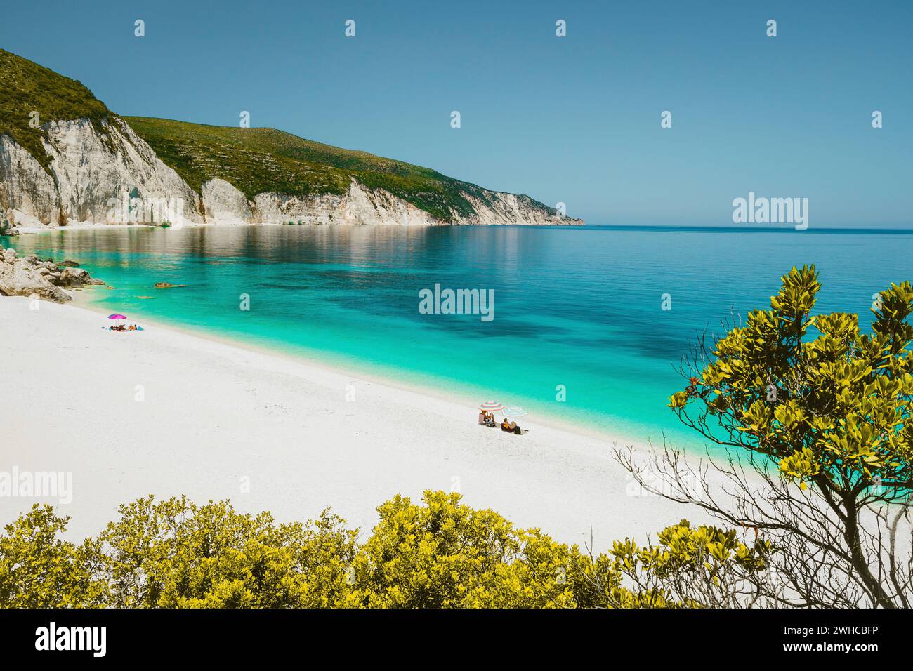Fantastische Lagune am Strand von Fteri, Cephalonia Kefalonia, Griechenland. Touristen entspannen sich unter dem Sonnenschirm in der Nähe des klaren blauen smaragdtürkisfarbenen Meerwassers. Weiße Steine im Hintergrund. Stockfoto