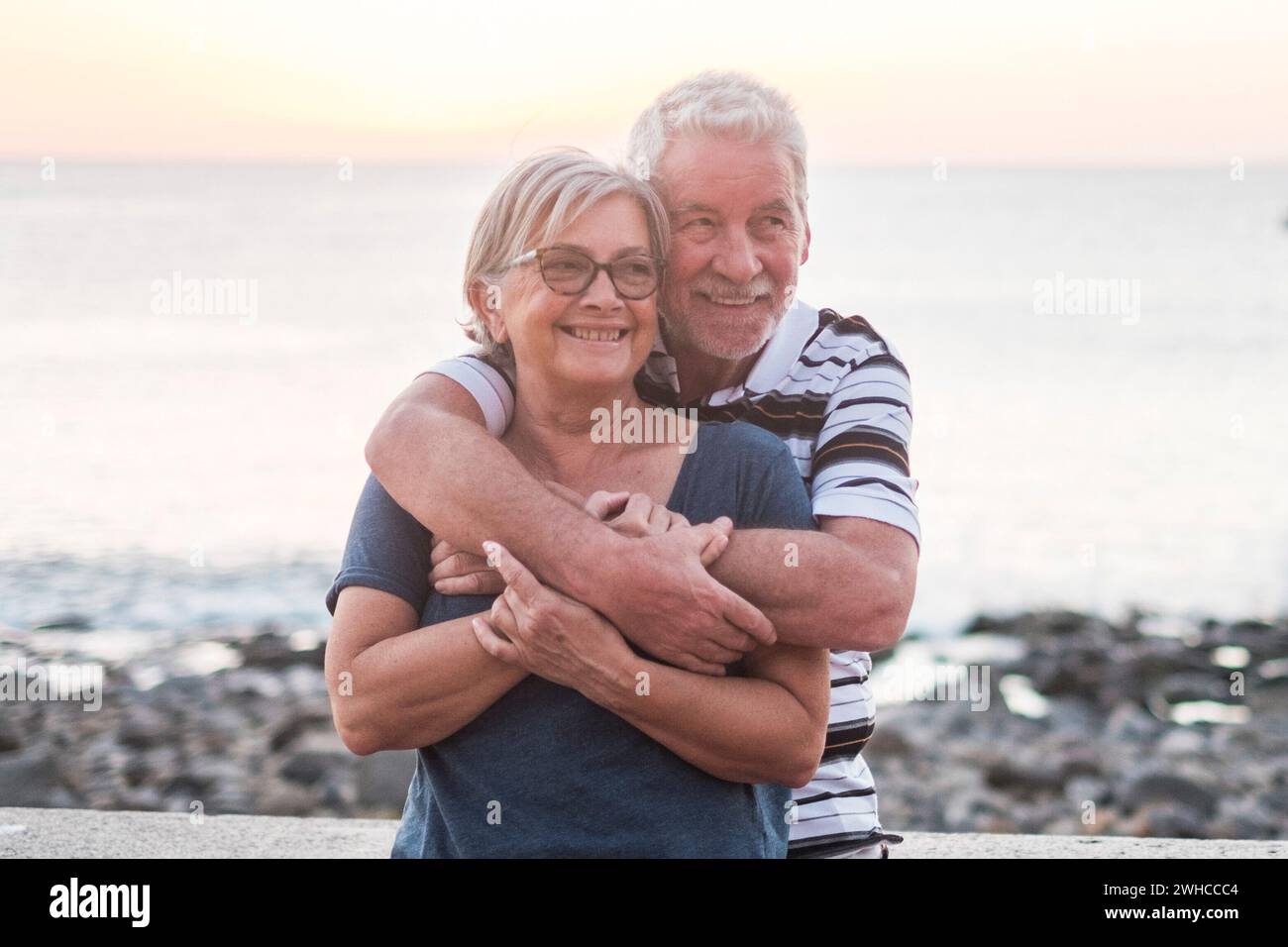 Ein paar Senioren, die mit viel am Strand hogging Von der Liebe - gemeinsam pensioniert - Frau mit Brille und Mann mit Meereshintergrund Stockfoto