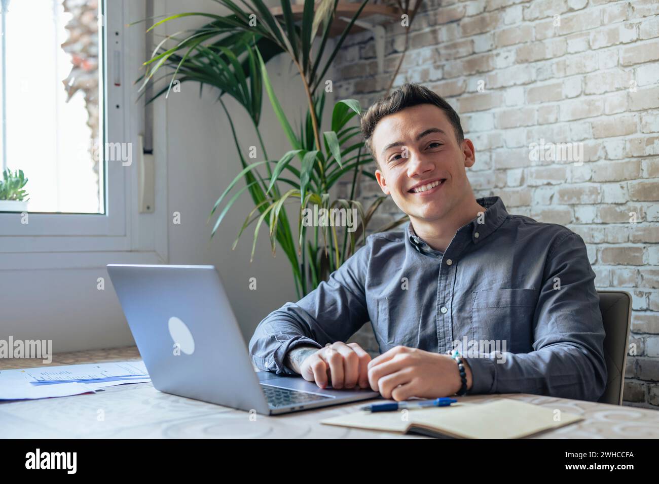 Gut aussehender Millennial Büroangestellter mit Brille am Schreibtisch vor dem Laptop und lächelnd in die Kamera. Erfolgreicher Mitarbeiter, Karrierefortschritt und Chancen, Eigentümer eines prosperierenden Geschäftskonzepts Stockfoto