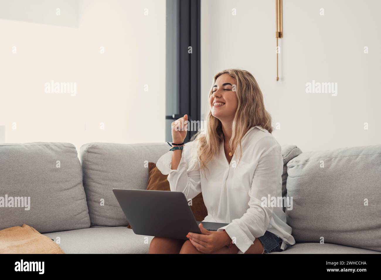 Eine junge glückliche Frau, die zu Hause mit einem Laptop auf der Couch feiert und Spaß hat. Euphorisches, fröhliches Mädchen, das Online-Preis genießt. Stockfoto