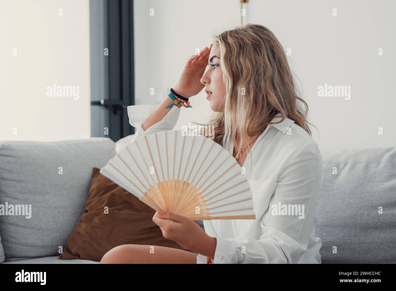 Überhitzte Frau, die am heißen Sommertag im Wohnzimmer auf der Couch sitzt und sich unwohl fühlt, leidet unter einem hitzewehenden weißen Ventilator, um sich abzukühlen, Mädchen schwitzt Wohnung ohne Klimaanlagenkonzept Stockfoto