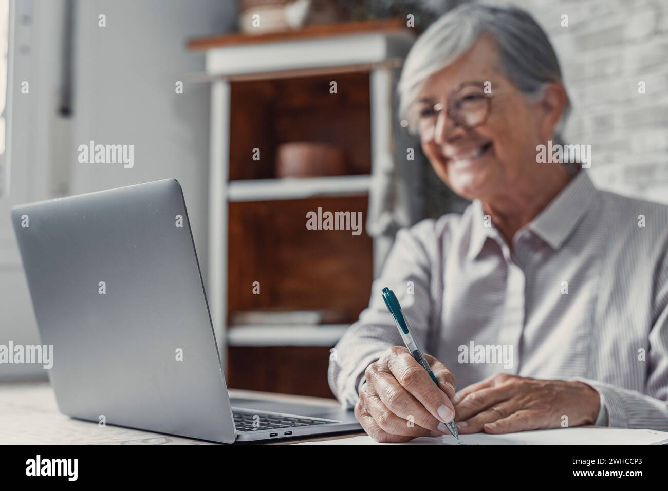 Veralteter Remote-Mitarbeiter. Konzentrierte Seniorin in Brille arbeitet am Laptop vom Heimbüro aus und liest elektronische E-Mail-Dokumente. Alte Frau Angestellte Freiberuflerin sitzt am Küchentisch durch pc-Tippbericht online Stockfoto