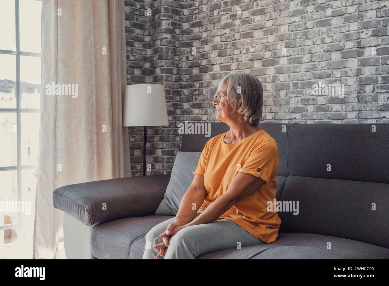 Positive nachdenkliche, hübsche blonde Frau mittleren Alters, die auf dem Sofa sitzt, die Hände am Kinn hält, mit guten Gedanken auf das Fenster blickt, über zukünftigen Ruhestand nachdenkt, plant, träumt Stockfoto