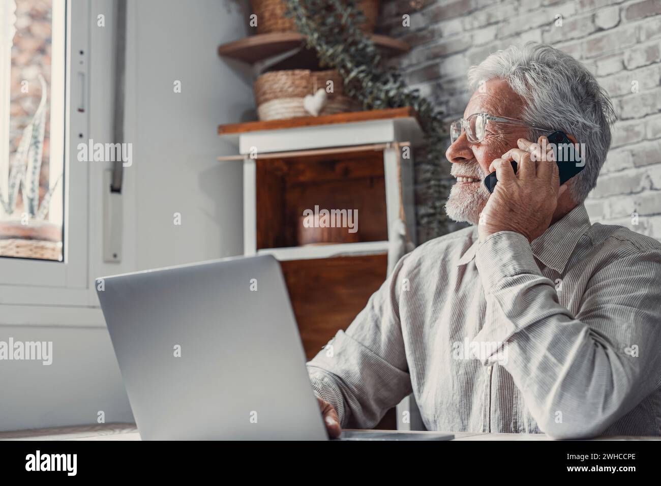 Porträt eines lächelnden Reifen Mannes sitzen am Tisch zu Hause reden über Handyarbeit auf Computer-Gadget. Smart Senior 70er-Männer haben Smartphone-Gesprächsgespräche, verwenden Sie Laptop. Ältere Menschen und Technologiekonzept. Stockfoto