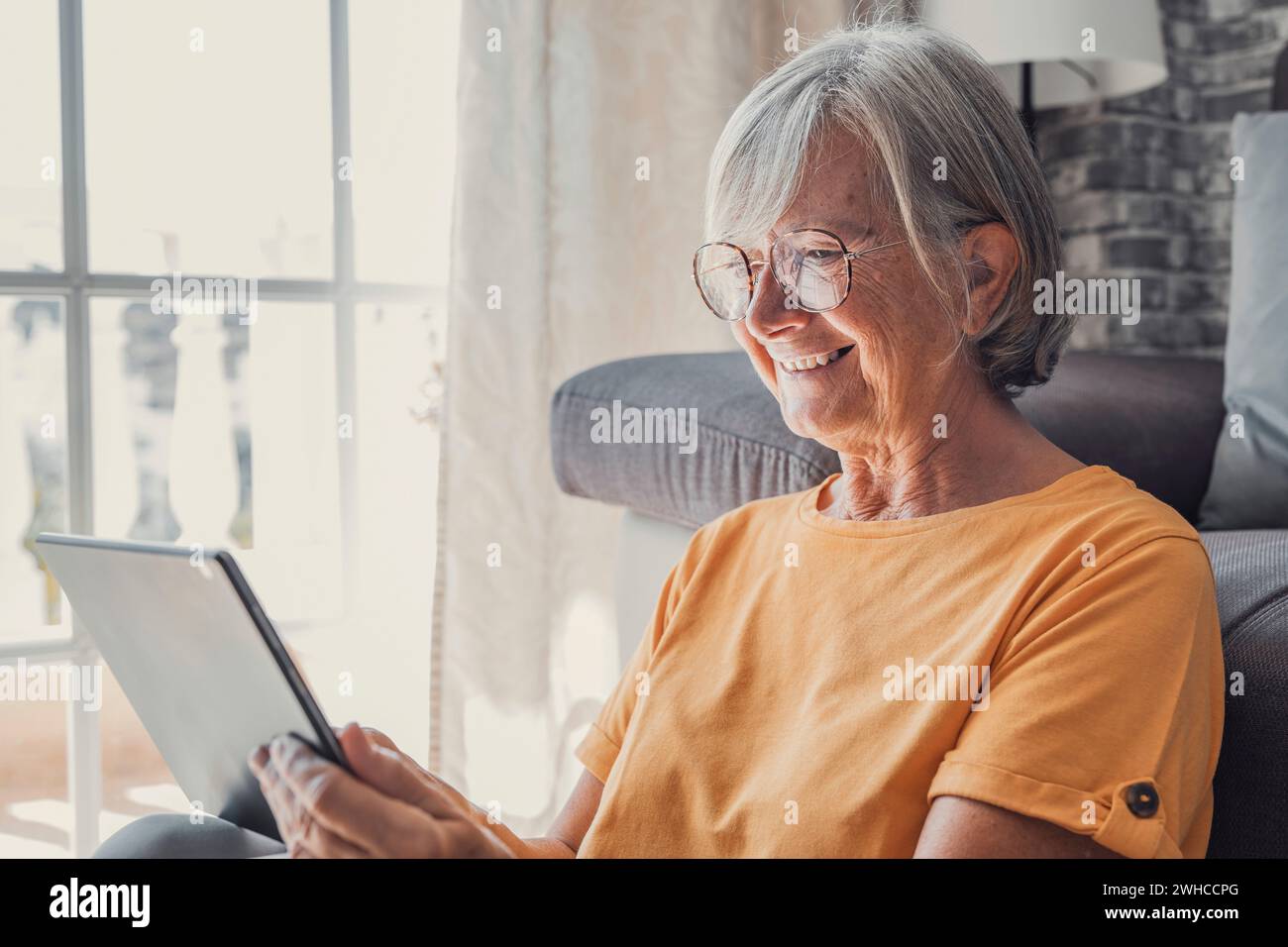 Lächelnde weiße Frau mittleren Alters sitzt auf der Couch im Wohnzimmer und surft im drahtlosen Internet auf einem Tablet, glückliche moderne ältere Frau entspannen sich auf dem Boden zu Hause mit Pad-Gerät, älteres Technologiekonzept Stockfoto