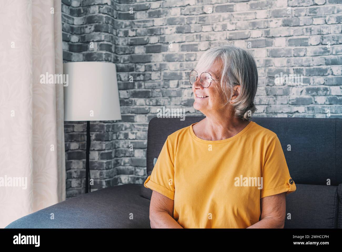 Positive nachdenkliche, hübsche blonde Frau mittleren Alters, die auf dem Sofa sitzt, die Hände am Kinn hält, mit guten Gedanken auf das Fenster blickt, über zukünftigen Ruhestand nachdenkt, plant, träumt Stockfoto