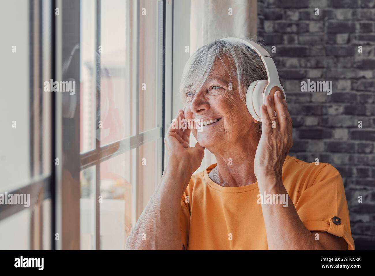 Seitenansicht glückliche alte, reife Rentnerin, die populäre Musik in Kopfhörern hört, sich alleine entspannen und ein Gedankenfenster aussah, friedliche, unbeschwerte Wochenendzeit allein zu Hause genießen, stressfreie Freizeitbeschäftigung. Stockfoto
