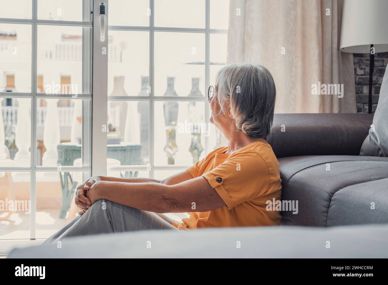 Positive nachdenkliche, hübsche blonde Frau mittleren Alters, die auf dem Sofa sitzt, die Hände am Kinn hält, mit guten Gedanken auf das Fenster blickt, über zukünftigen Ruhestand nachdenkt, plant, träumt Stockfoto