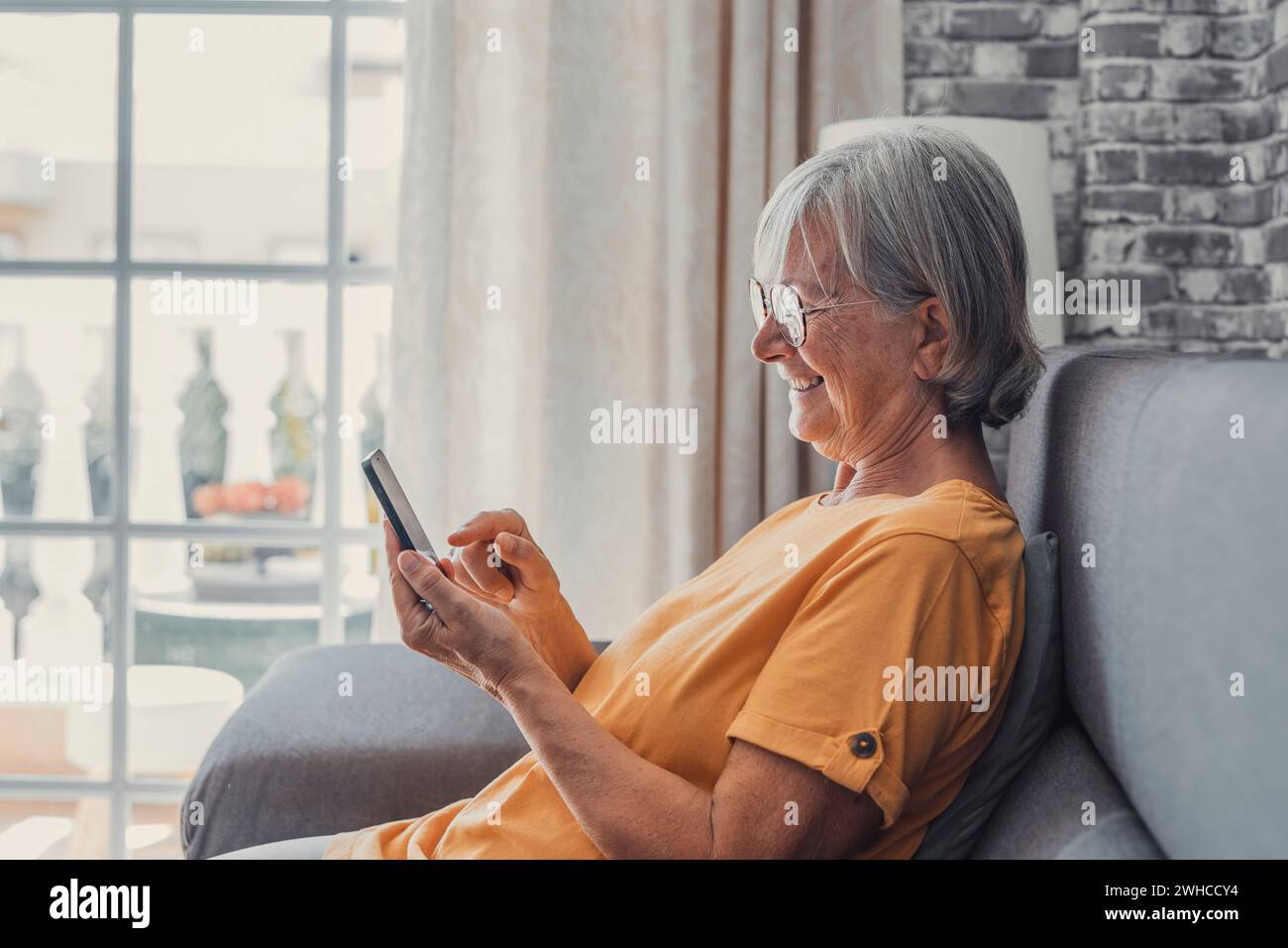 Lächelnde weiße Frau mittleren Alters sitzt neben der Couch im Wohnzimmer und surft drahtloses Internet am Telefon, glückliche moderne ältere Frau entspannen sich auf dem Boden zu Hause mit Smartphone-Gerät, älteres Technologiekonzept Stockfoto