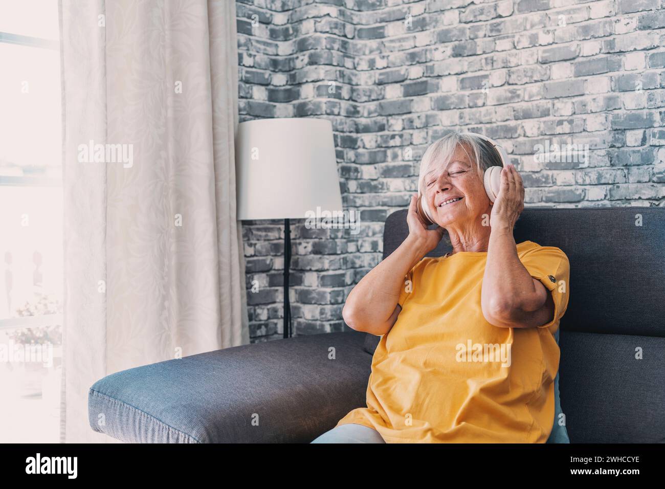 Seitenansicht glückliche alte, reife pensionierte Frau, die populäre Musik in Kopfhörern hört, sich allein auf einem gemütlichen Sofa entspannt, friedliche, unbeschwerte Wochenendzeit allein zu Hause genießt, stressfreie Freizeitbeschäftigung. Stockfoto