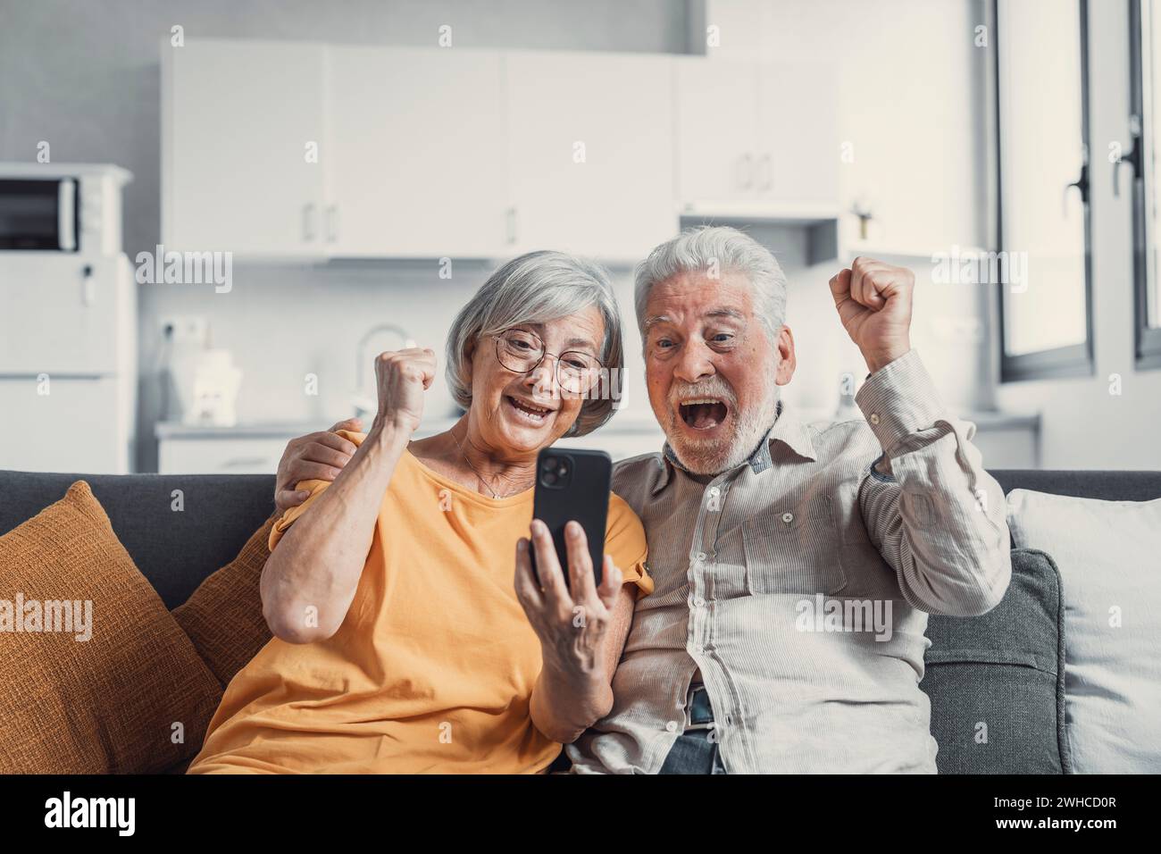 Glückliches älteres Paar sitzt auf der Couch und starrt auf den Handybildschirm und sieht überglücklich aus, schreien vor Freude, lesen fantastische Nachrichten, erhalten ein tolles kommerzielles Angebot, erhöhen die Rente. Erfolg, Leistungskonzept Stockfoto