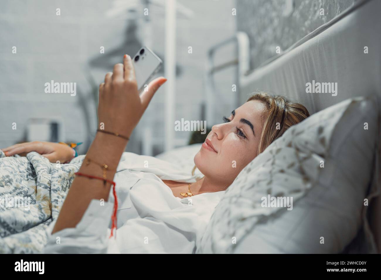 Gadget-sucht. Ruhig lächelnde junge Frau oder Mädchen im Teenageralter, die an einem gemütlichen Bett liegt, das Handy in der Hand hält und auf den Bildschirm schaut, um den Account des sozialen Netzwerks zu überprüfen, bevor sie einschlafen oder nach dem Aufwachen Stockfoto