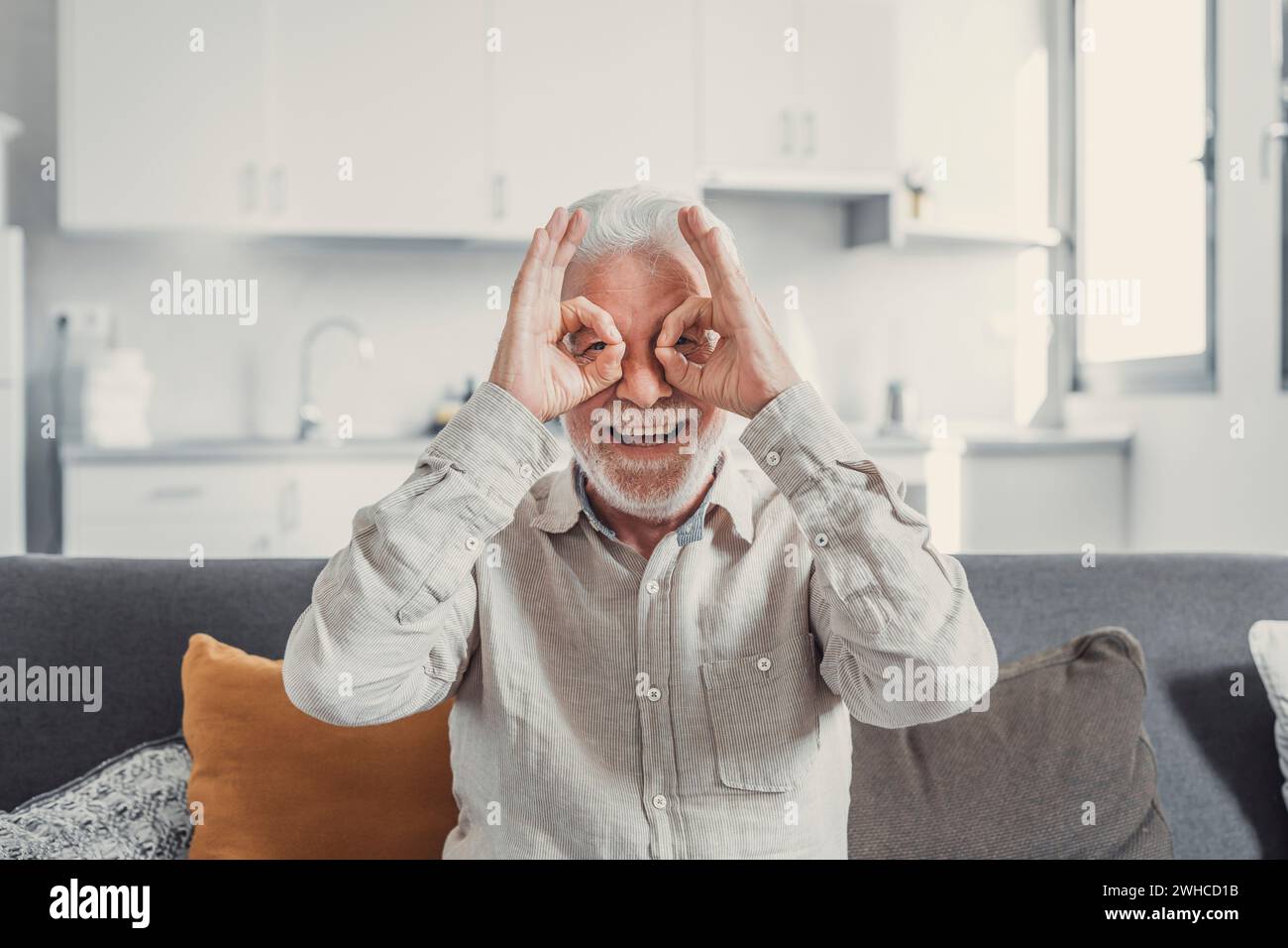 Kopffoto und Porträt eines alten, reifen Mannes, der auf dem Sofa sitzt und in die Kamera schaut und lustige Gesichter mit lächelnden und lachenden Fingern und Händen macht. Gesunde Senioren genießen zu Hause Stockfoto