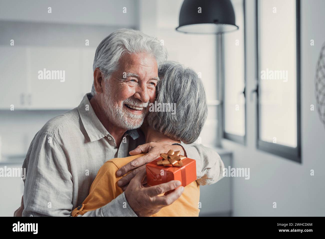Liebevoll lächelnde ältere Frau schenkt dem lächelnden alten Mann der 80er Jahre eine Geschenkbox und gratuliert zum Geburtstag oder Hochzeitstag. Fröhlicher Seniorenmann, der aufgeregt ist, von seiner Frau ein Geschenk zu bekommen Stockfoto