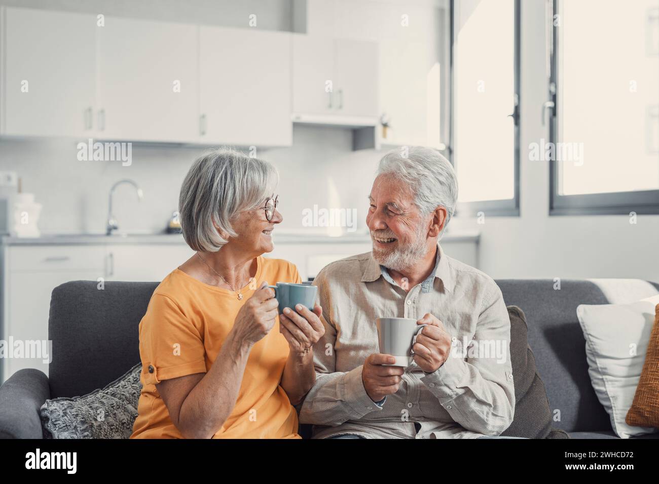 Glückliche, reife 50er Jahre Mann und Frau sitzen auf einem komfortablen Sofa im Wohnzimmer Genießen Sie Tee unterhalten, lächelnde ältere 60er Jahre Paar entspannen sich auf der Couch zu Hause trinken Kaffee plaudern Lachen am Freizeit-Wochenende Stockfoto