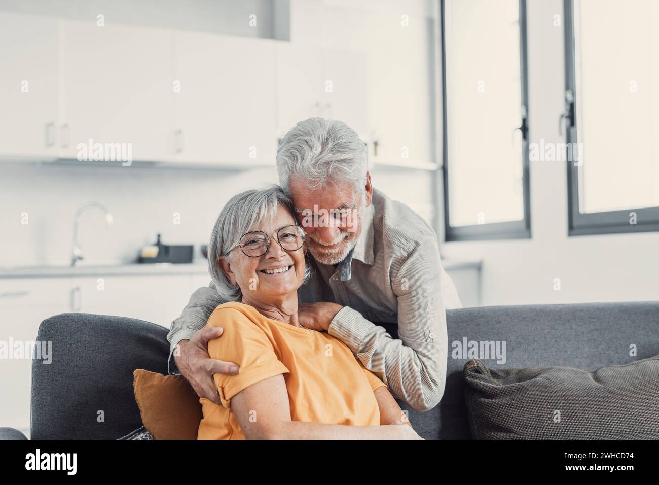 Kopffoto Porträt lächelnder älterer 60er Jahre Mann und Frau sitzen auf der Couch, umarmen Kuscheln, glückliches reifes altes Paar ruhen auf Sofa im Wohnzimmer umarmen Blick auf Kamera zeigen Liebe und Sorgfalt Stockfoto