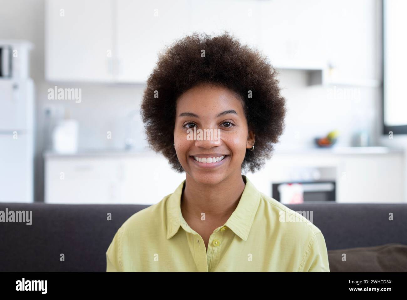 Porträt eines glücklichen Millennials-Schwarzen, das zu Hause auf dem Sofa sitzt und mit einem zahnlosen Lächeln in die Kamera blickt. Schöne afroamerikanische junge Frau, weibliches Model im Haus. Kopfschuss Stockfoto