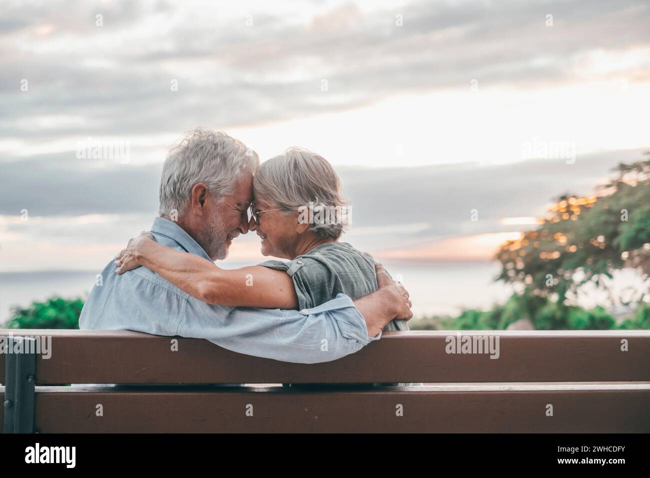 Kopfaufnahme Nahaufnahme Porträt glückliche grauhaarige Frau mittleren Alters, die sich an einen lächelnden älteren Ehemann kuschelt, genießt es, auf einer Bank im Park zu sitzen. Ein liebevolles, altes Familienpaar, das den Sonnenuntergang umschließt. Stockfoto
