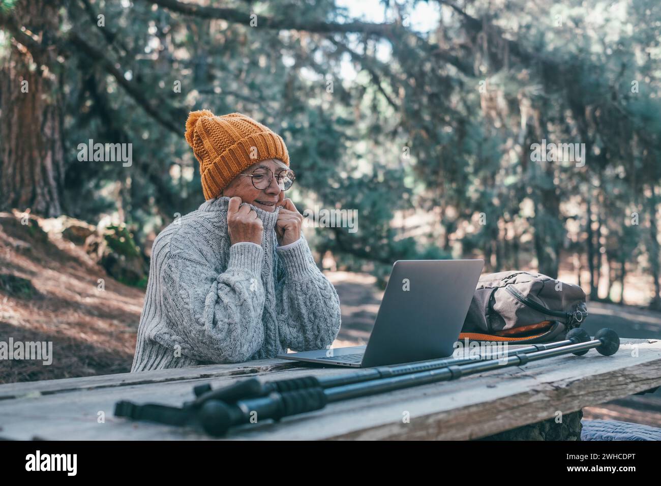 Porträt, Nahaufnahme einer niedlichen Person des alten mittleren Alters, die im Freien einen Computer benutzt und an einem Holztisch im Wald des Berges in der Natur sitzt, mit Bäumen um sie herum. Stockfoto